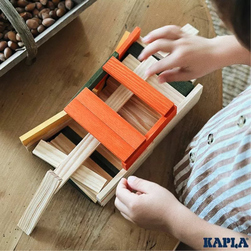 Child's hands engaging in open-ended play, assembling a colorful wooden structure with KAPLA® 200 Autumn Box planks on a wooden table, scattered with hazelnuts.