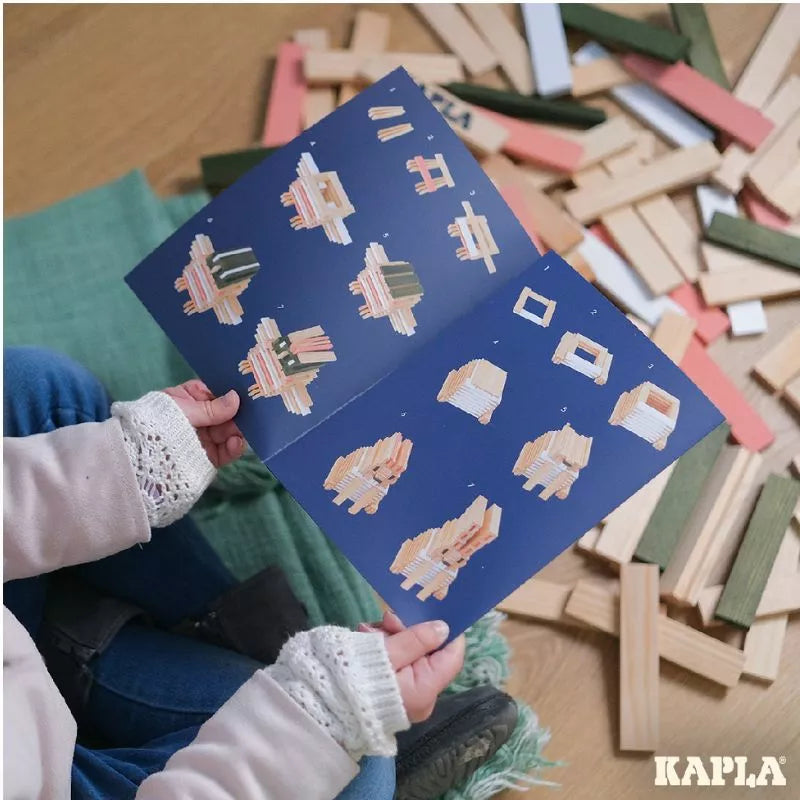 A child is holding a book with pictures of animals on it, while playing with construction toys made of KAPLA® 200 Spring Box planks.