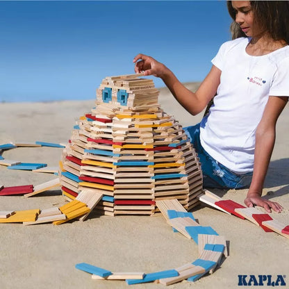 A focused individual carefully places a wooden plank on an intricate dome structure made of colorful KAPLA® 200 Summer Box planks on a sandy background, demonstrating the joy of open-ended play with this construction toy.