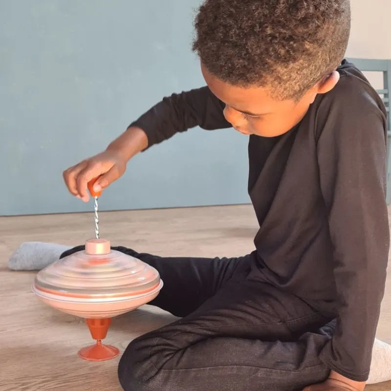 A child in a black long-sleeve shirt and pants is sitting on a wooden floor, intently focused on the Large Spinning Top Countryside—a large red and silver sensory toy with an orange handle.