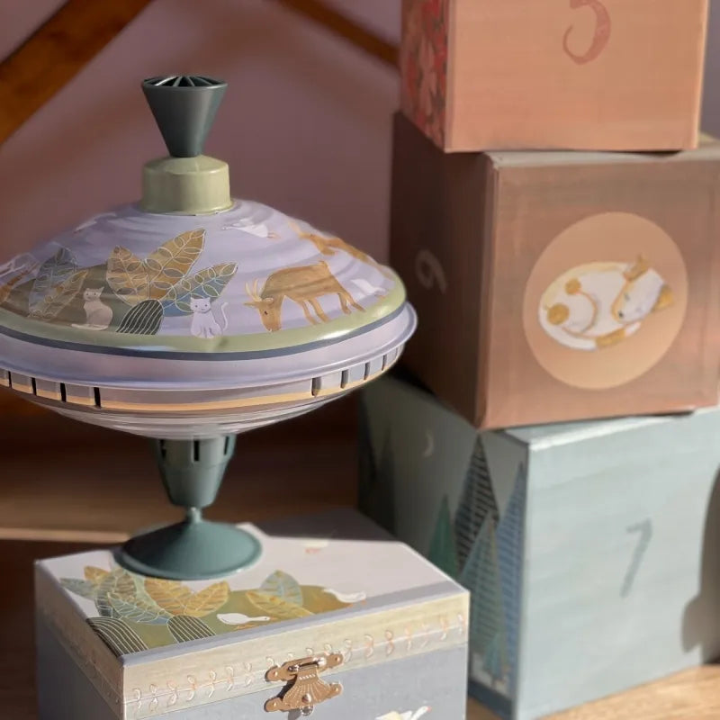 The Large Spinning Top Countryside, adorned with woodland animal motifs, rests on a wooden surface. Surrounding it are pastel-colored blocks and boxes, each block displaying a number and an animal illustration. This charming sensory scene is gently bathed in sunlight.