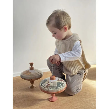 A young child kneels on a wooden floor, playing with the Large Spinning Top Noah’s Ark. The child wears a beige knit vest over a white long-sleeve shirt and light-colored pants. This nostalgic toy complements the scene, set against a plain white wall.