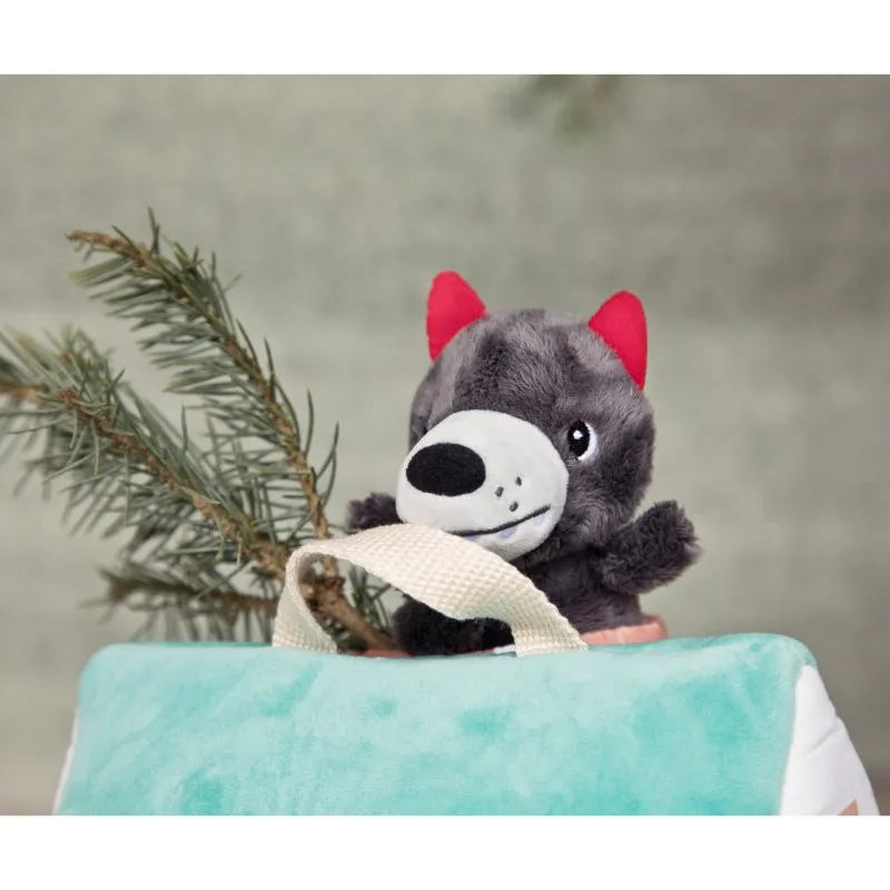 A small gray plush wolf with red ears peeks out from behind the blue and white fabric house of the "Lilliputiens Houses Of The Wolf And The 3 Little Pigs," perfect for creative play. A sprig of green pine needles is visible to the left of the wolf. The background is soft and out of focus, enhancing fine motor skills as children interact with this charming fabric toy.