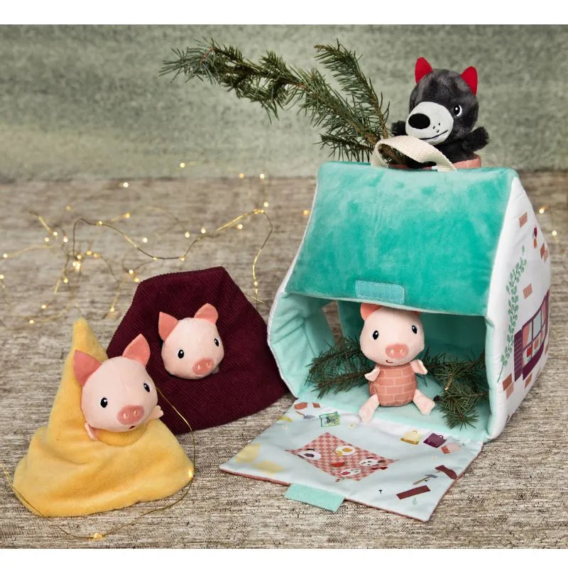 The Lilliputiens Houses Of The Wolf And The 3 Little Pigs set features three plush pigs and a wolf, with soft accessories and a playhouse—two pigs in cozy covers, one inside the house, and the wolf peeking over the roof with a branch.