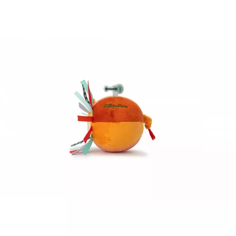 A Lilliputiens Jack Ball Lion plush orange sensory toy with colorful ribbon tags and a metal key sticking out of its top, set against a white background.