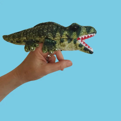 A hand holds The Puppet Company Alligator Finger Puppet – Little Creatures Range, a green and yellow soft puppet with an open mouth and white teeth, against a blue background. Ideal for playful storytelling.