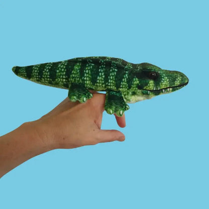 A hand holds The Puppet Company Crocodile Finger Puppet from the Little Creatures Range, showing its green patterned design against a plain light blue background.