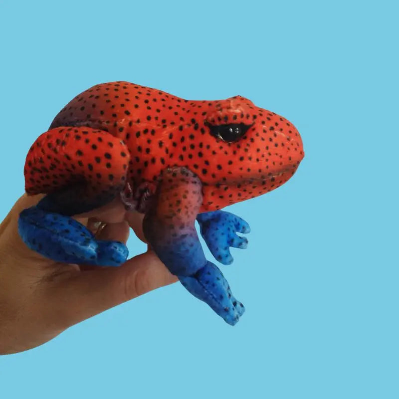 A hand displays The Puppet Company Red Frog Finger Puppet from the Little Creatures Range, featuring a vibrant red body with black spots and blue legs, ideal for imaginative play, set against a plain light blue background.