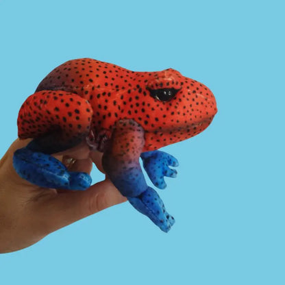 A hand displays The Puppet Company Red Frog Finger Puppet from the Little Creatures Range, featuring a vibrant red body with black spots and blue legs, ideal for imaginative play, set against a plain light blue background.