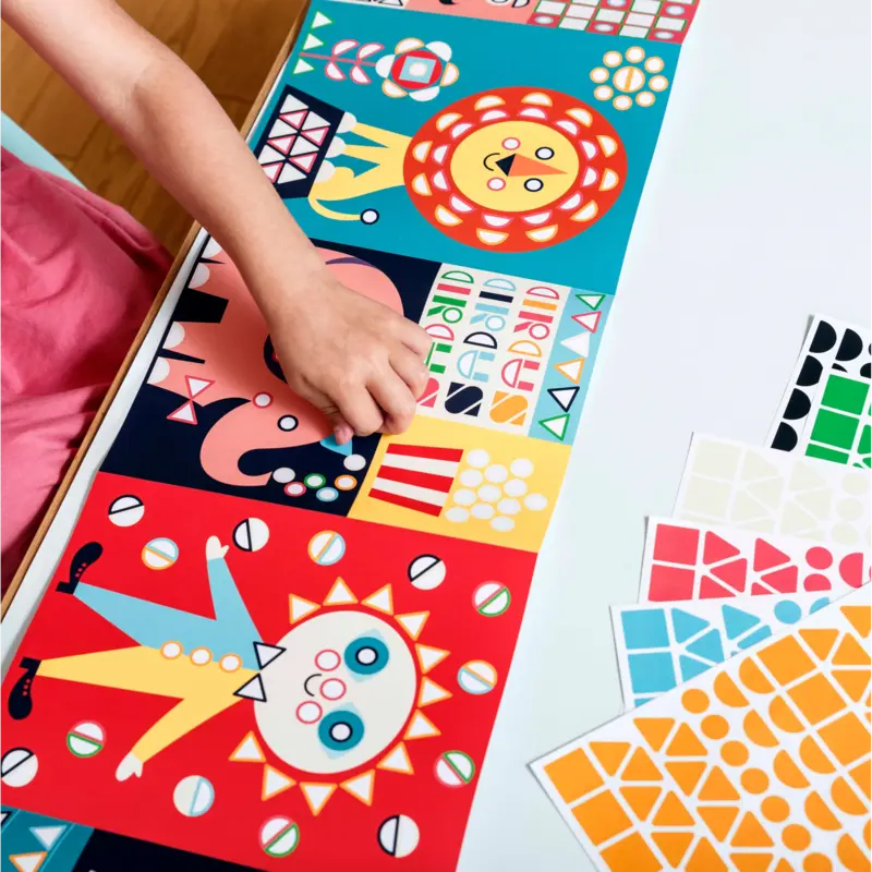 A child enjoys Poppik Sticker Panorama – Circus, arranging colorful geometric stickers onto illustrated circus animal scenes, like a lion and elephant, while more sticker sheets and a fold-out panoramic poster are nearby.