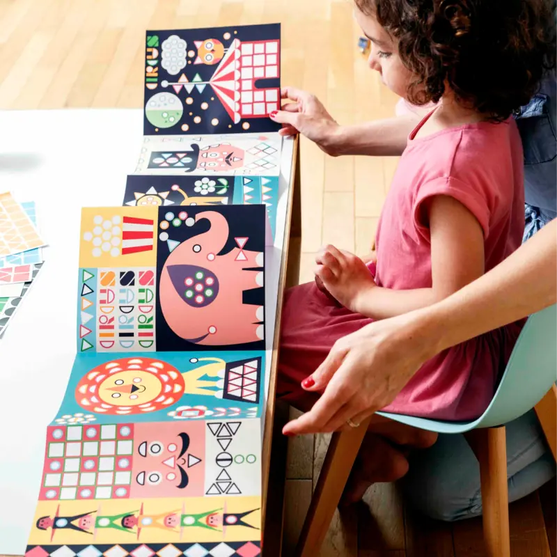 A child in a pink dress sits at a table with an adult, enjoying the Poppik Sticker Panorama – Circus activity as they arrange colorful geometric animal sticker pieces in a row, making creative fun for children.
