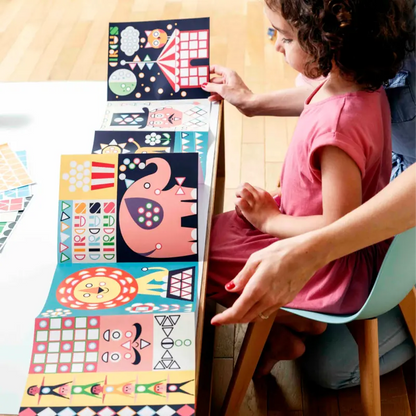A child in a pink dress sits at a table with an adult, enjoying the Poppik Sticker Panorama – Circus activity as they arrange colorful geometric animal sticker pieces in a row, making creative fun for children.