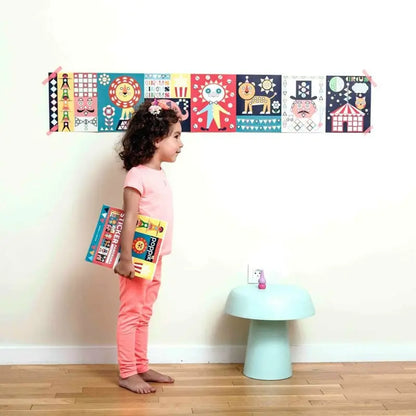 A young girl stands barefoot on a wooden floor, holding colorful books. She faces a wall with the Poppik Sticker Panorama – Circus displayed above a small, light blue mushroom-shaped stool.