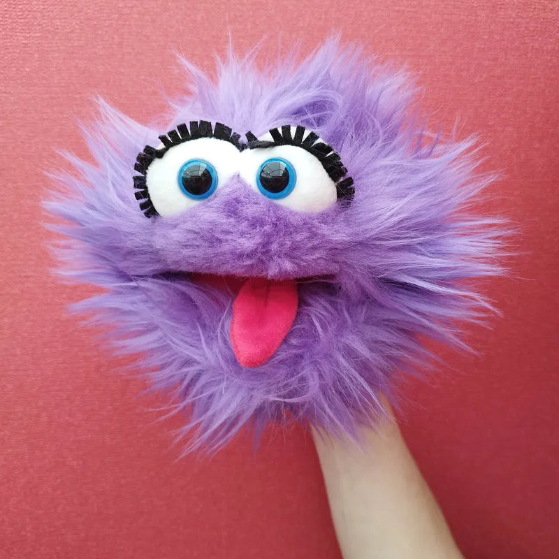 The Living Puppets Flob Hand Puppet features a fluffy, purple exterior, large blue eyes with black eyelashes, and a wide mouth with a pink tongue sticking out. Perfect for creative play, this delightful puppet is set against a bright pink wall.