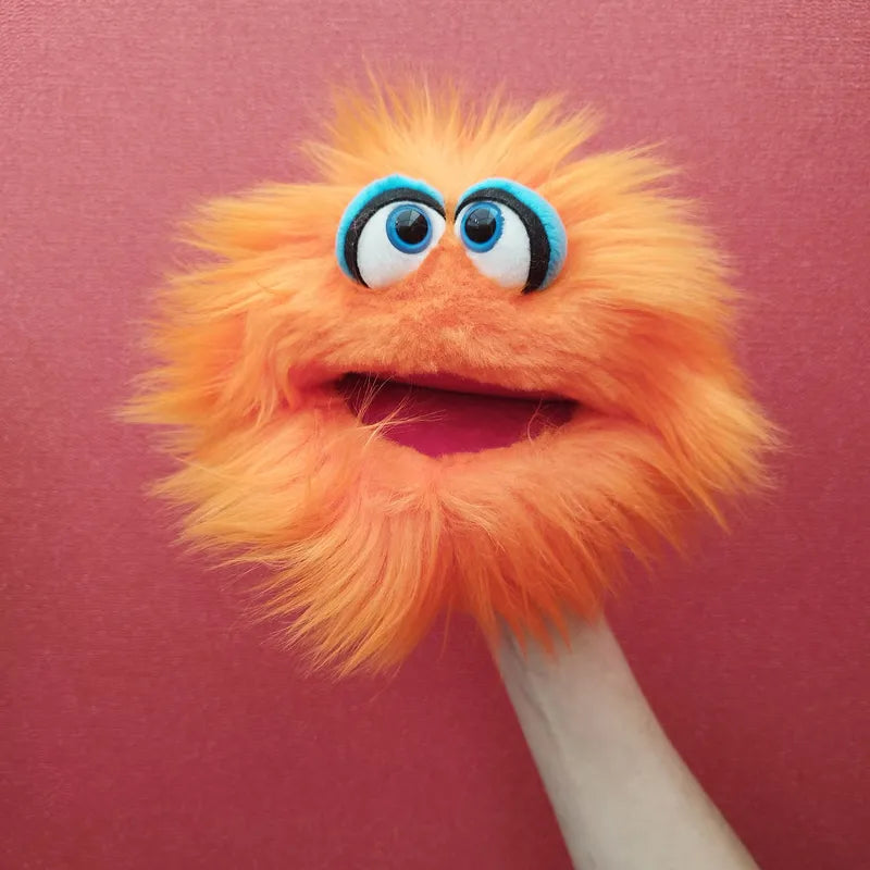 The Living Puppets Klops Hand Puppet, featuring bright orange fur, large blue eyes with black pupils, and a big open mouth, is set against a pinkish-red background. The puppet's mouth moves as if engaging in creative play, animated by a hand inside it.