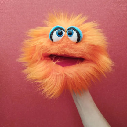 The Living Puppets Klops Hand Puppet, featuring bright orange fur, large blue eyes with black pupils, and a big open mouth, is set against a pinkish-red background. The puppet's mouth moves as if engaging in creative play, animated by a hand inside it.