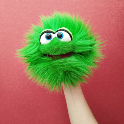 A Living Puppets Blappermouth Hand Puppet, featuring a bright green color and fuzzy texture, big googly eyes, and a red mouth, is being held up against a textured red background. The puppet's expressive eyes give it a lively appearance, making it perfect for creative play.