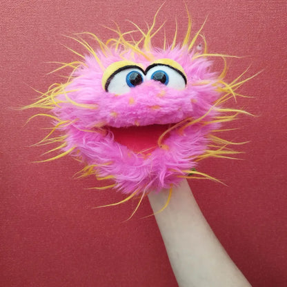 The Living Puppets Monster Hand Puppet Knaller, resembling a fuzzy pink creature with yellow and orange accents, is shown against a reddish background. The puppet has large blue eyes and an open mouth, giving it an expressive, animated appearance. The arm controlling the hand puppet is visible.