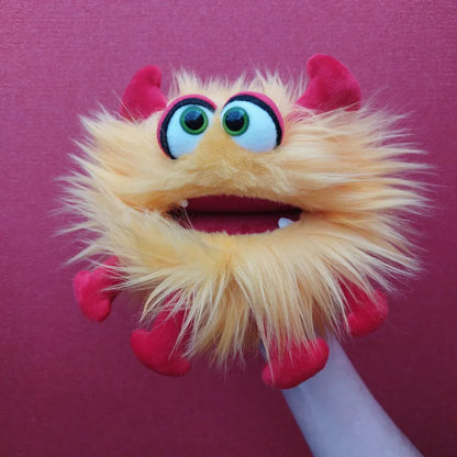 Against a bright red background, the Living Puppets Monster Hand Puppet Gisa is displayed in all its vibrant glory, showcasing its fuzzy yellow and orange features, big green eyes, wide open mouth with one protruding tooth, and red horns, paws, and feet.