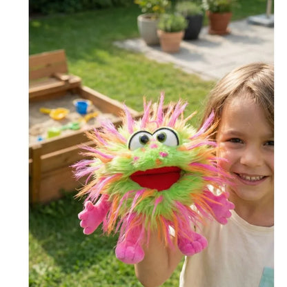 A smiling child holds the colorful Living Puppets Knibbels Hand Puppet, featuring green and pink fur, big eyes, and an open red mouth. They play near a wooden sandbox on green grass in a garden—perfect for creative fun.