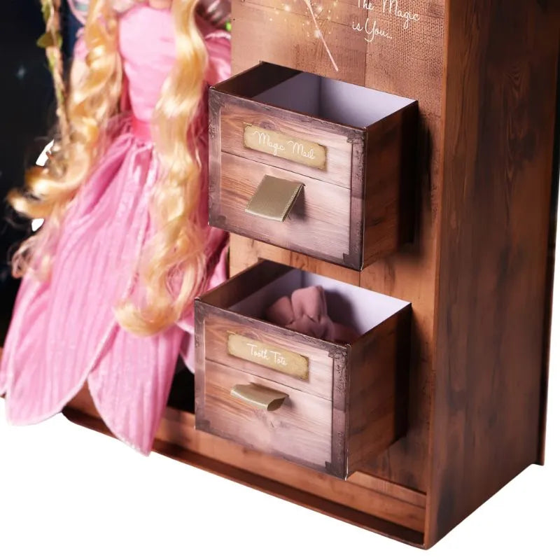 The My Forever Tooth Fairy Gift Set includes a charming wooden storage unit with two small drawers labeled "Magic Mail" and "Troll Toll." Additionally, a doll wearing a pink dress with long blonde hair is partially visible to the left of the unit, sparking children's imagination to fill this keepsake Tooth Fairy Gift Box.