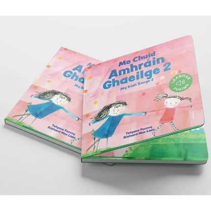 Two copies of "Mo Chuid Amhráin Ghaeilge 2" are stacked, showing playful drawings of two girls holding hands on a pink and green cover—an ideal book for introducing Irish songs to children.