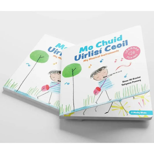 Two illustrated "Mo Chuid Uirlisí Ceoil" children's books show a cartoon child playing a drum and making music, with grass and a tree on the cover. The books are stacked on a light surface.