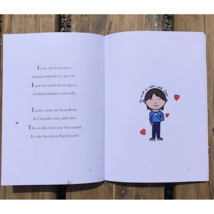 An open children's book is laying on a wooden surface. The left page contains an inspirational poem about practicing mindfulness and calmness. The right page has a simple illustration of a person softly smiling with closed eyes, hands on their heart, surrounded by small hearts, radiating self-compassion.