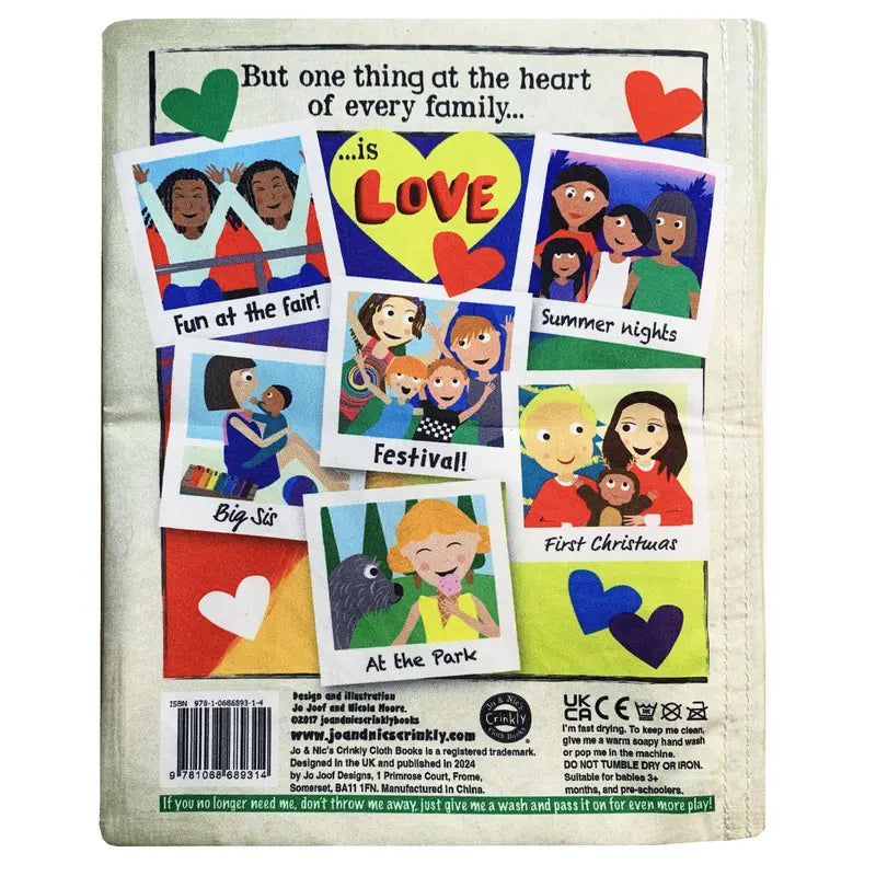 The image shows the back cover of the "Crinkly Cloth Book What Makes A Family," featuring colorful cartoons of diverse families at fairs, festivals, parks, and Christmas. The text reads: “But one thing at the heart of every family... is LOVE.”.