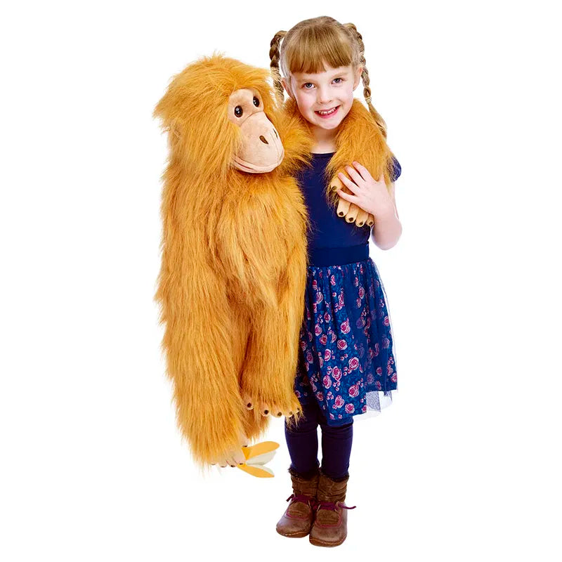 A young girl with braided hair and a blue dress smiles while holding The Puppet Company Large Puppet Orangutan. The plush toy features a light brown hue and long arms, set against a plain white background.