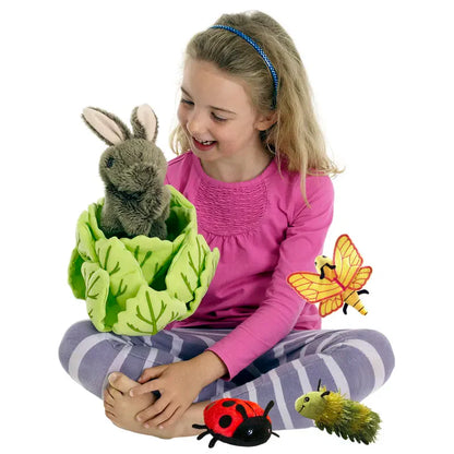 A smiling girl in a pink shirt and striped leggings sits cross-legged, holding The Puppet Company Hide-Away Rabbit plush in a cabbage, with butterfly, ladybug, and caterpillar plush insects on her lap and colorful finger puppets nearby.