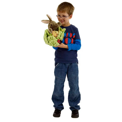 A young boy in glasses, a blue shirt, and jeans smiles while holding The Puppet Company Hide-Away Rabbit and a green basket with toy vegetables. The background is plain white.