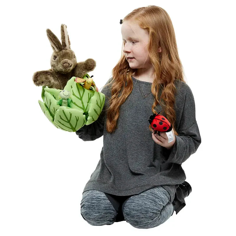 A young girl with long red hair kneels, smiling as she holds The Puppet Company Hide-Away Rabbit, a soft glove puppet, along with frog and ladybug plush toys.