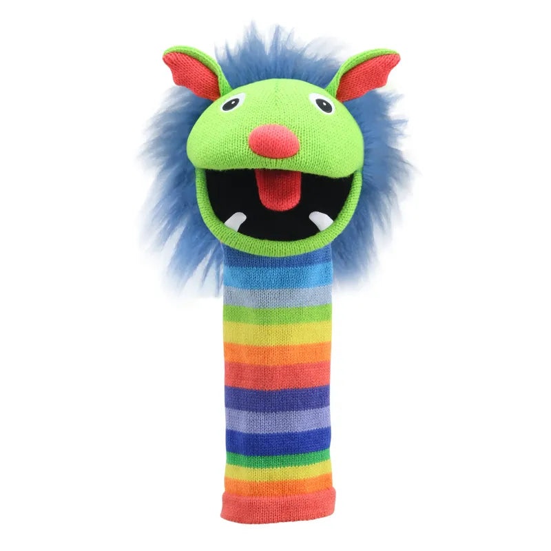 The Puppet Company Sockette Puppet Rainbow is a vibrant hand puppet featuring a green head, blue fur hair, and a red tongue with an integrated squeaker. It has big white eyes with black pupils, pink ears, and two small white teeth sticking out of its mouth. The body showcases rainbow-striped knitting, with colors spanning from red to purple.