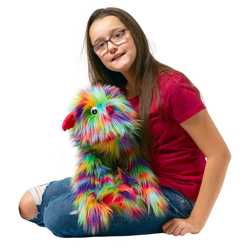 A girl in glasses and a red shirt sits cross-legged, holding The Puppet Company Rainbow Monster puppet with rainbow fur, featuring a squeaker and red accents. She smiles at the camera against a white background.