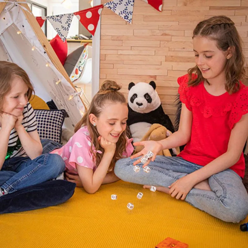 Three girls playing Rory's Story Cube Classic on a yellow mat in a cozy room with a teepee and plush toys in the background.