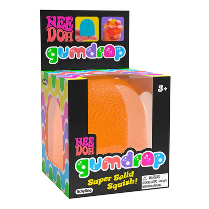 A packaged Gumdrop Needoh fidget toy displays a bright orange, textured squishy ball through a clear window. The colorful box claims "Super solid Squish!" and is ideal for stress relief for ages 3 and up.