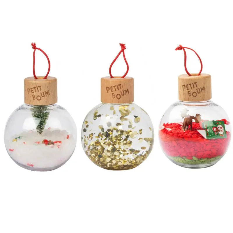 Christmas Sensory Baubles: three round, clear ornaments with wooden "PETIT BOUM" caps. Each features festive decorations—a snowy tree, gold glitter, red beads with a reindeer and gift box—and red hanging loops for little hands.