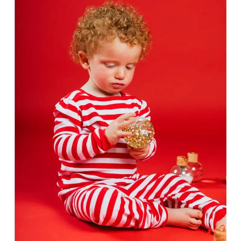 A young child in red and white striped pajamas sits on the floor against a red background, looking intently at a Christmas Sensory Bauble filled with gold glitter in their hands.