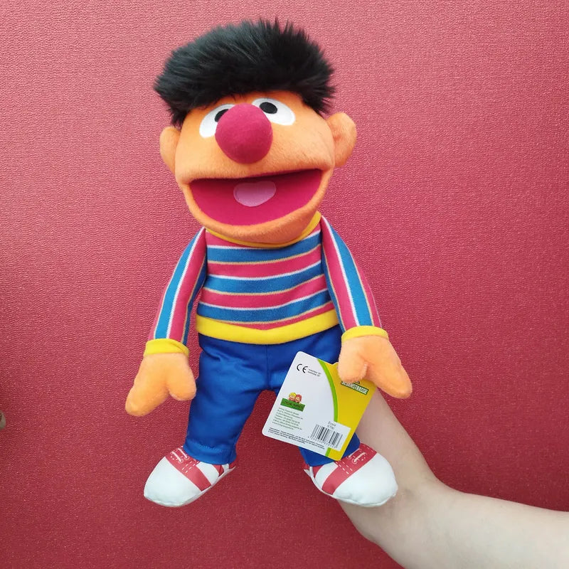 A hand holds up the colorful Living Puppets Ernie Hand Puppet 33cm with black hair, a red round nose, and a wide open mouth. The Ernie toy wears a striped long-sleeve shirt, blue pants, and red-and-white sneakers. The background is solid red. A tag is attached to the puppet’s left hand.