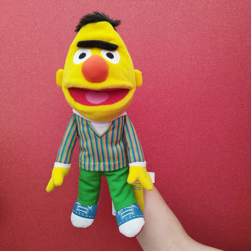 A hand is holding the Living Puppets Bert Hand Puppet 33cm against a red background. The puppet has a yellow face, bushy black eyebrows, an orange nose, and is wearing a striped shirt with green pants and blue shoes.