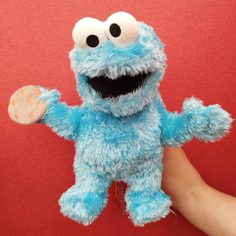 A hand is holding up a Living Puppets Cookie Monster Hand Puppet, with its blue fur, large white eyes, and open mouth. The Sesame Street puppet holds a small cookie in one hand and is set against a vibrant red background.