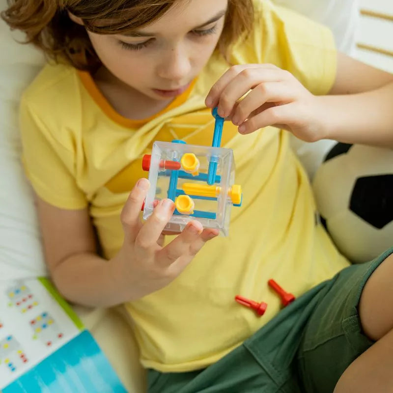 A young girl is playing with a SmartGames Criss Cross Cube while lying on a bed, indulging in logic and brainteasers.