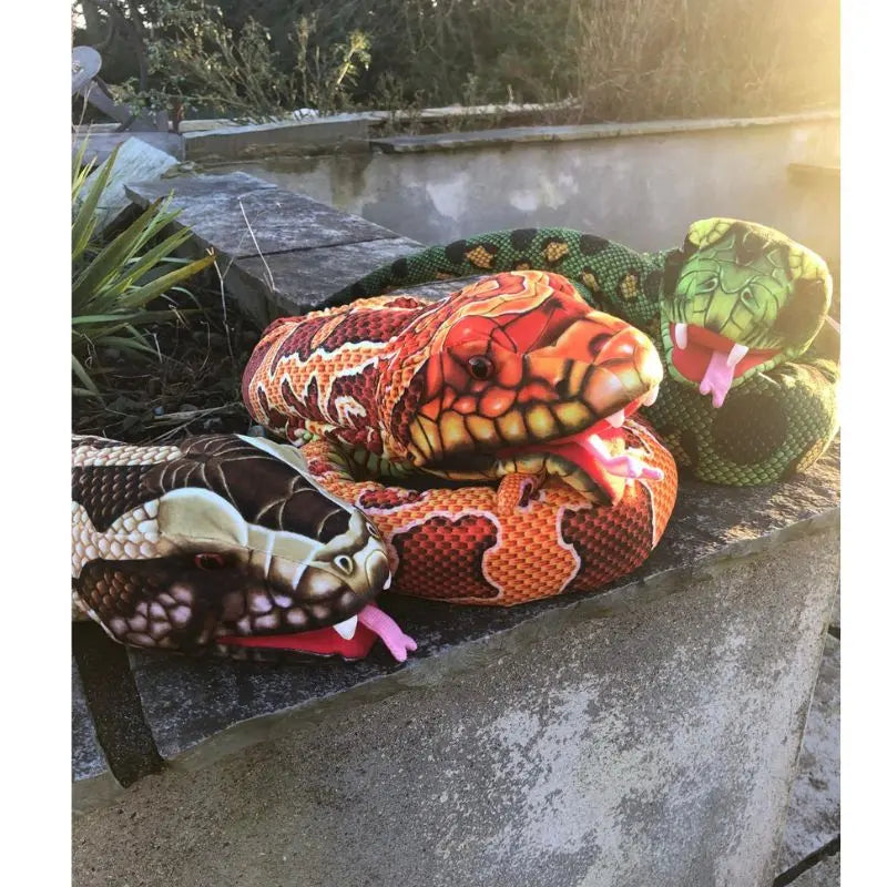 Three The Puppet Company Large Creature Snake plush toys—brown, orange, and green—rest outdoors on a concrete ledge with greenery and sunlight, inviting creative play and curiosity about nature.