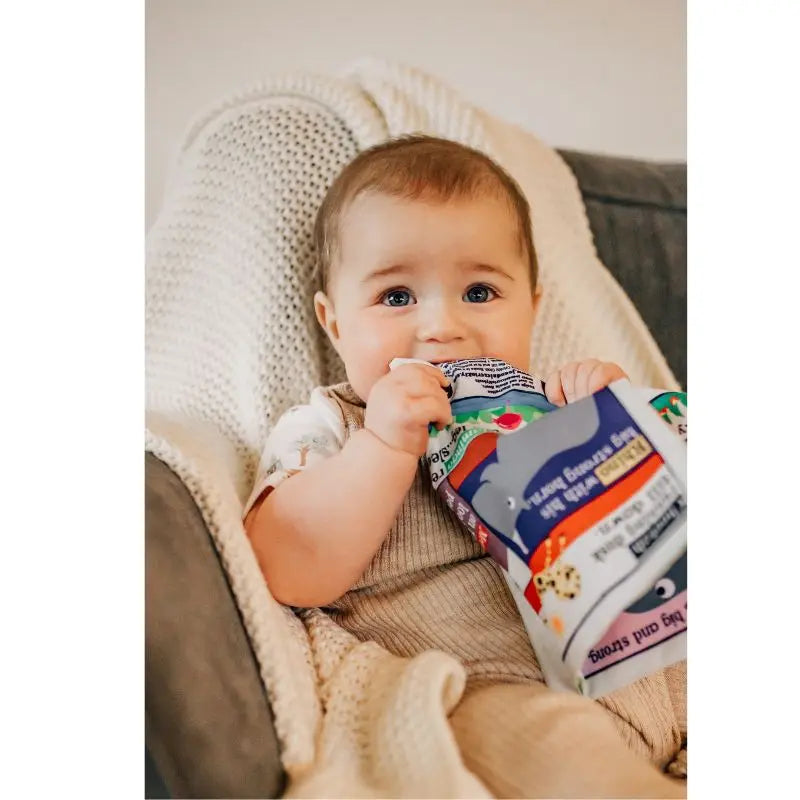 A baby with light brown hair and blue eyes sits on a cushioned chair, chewing on a colorful food pouch. Next to them is the Crinkly Cloth Book Count To 10 In German, adding to their sensory experience as they look curiously at the camera.