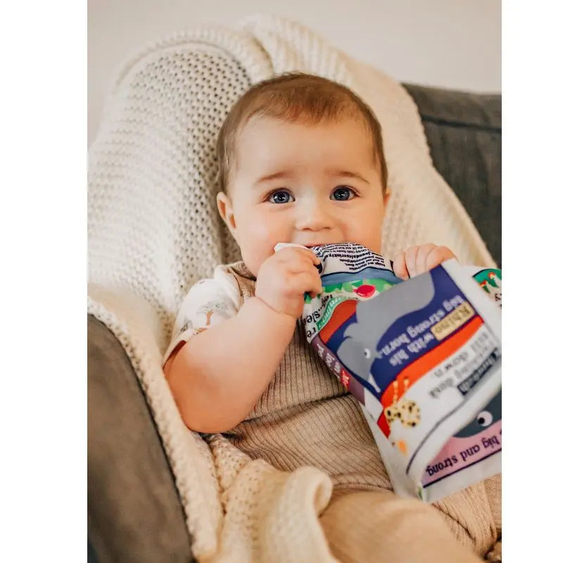 A baby with light brown hair sits in a gray armchair, holding and chewing on the Crinkly Cloth Book Count To 10 In Ukrainian with colorful animal illustrations, and looks up with wide eyes.
