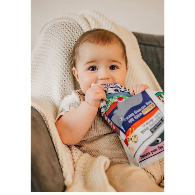A baby with blue eyes sits in a cozy chair, wrapped in a beige blanket, holding a colorful food pouch and sitting next to the Crinkly Cloth Book A-Z of Cats.