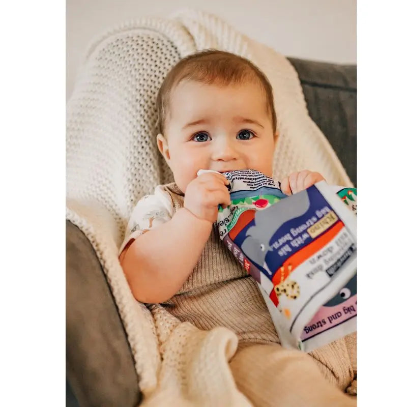 A baby with light brown hair and blue eyes sits on a cozy chair, wrapped in a beige blanket, holding the Crinkly Cloth Book Three Little Tales and gently chewing on it.