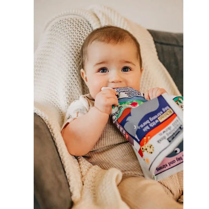 A baby with light brown hair and blue eyes sits on a cozy chair, wrapped in a beige blanket, holding the Crinkly Cloth Book Three Little Tales and gently chewing on it.