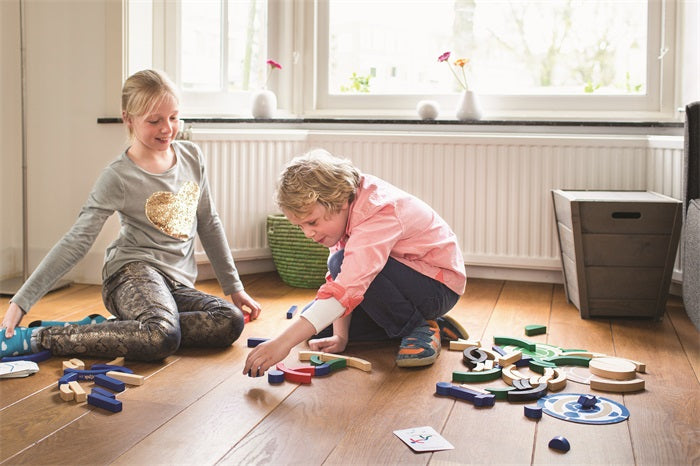 Two children play with the Buitenspeel Sport Blocks Puzzle Game on a wooden floor in a sunlit room. The girl smiles as she watches the boy, who is focused on solving the colorful puzzle, while sunlight pours in through large windows behind them.